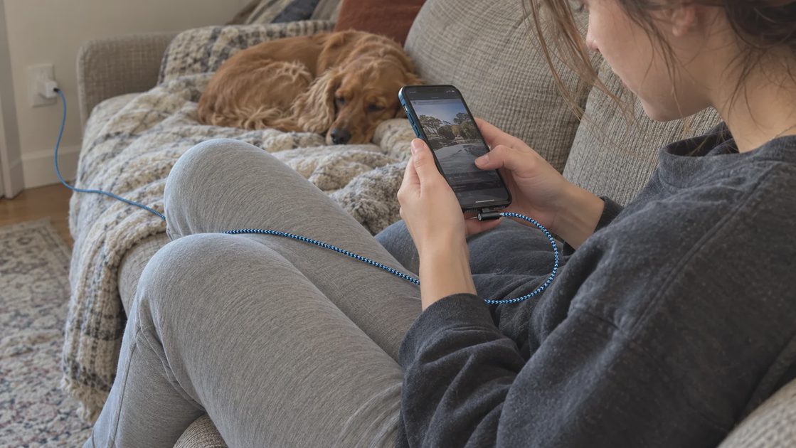Relaxed Sofa Charging with a 10ft Lightning Cable A woman sitting comfortably on a sofa uses an iPhone connected to a long blue braided right-angle cable, while a dog rests beside her.